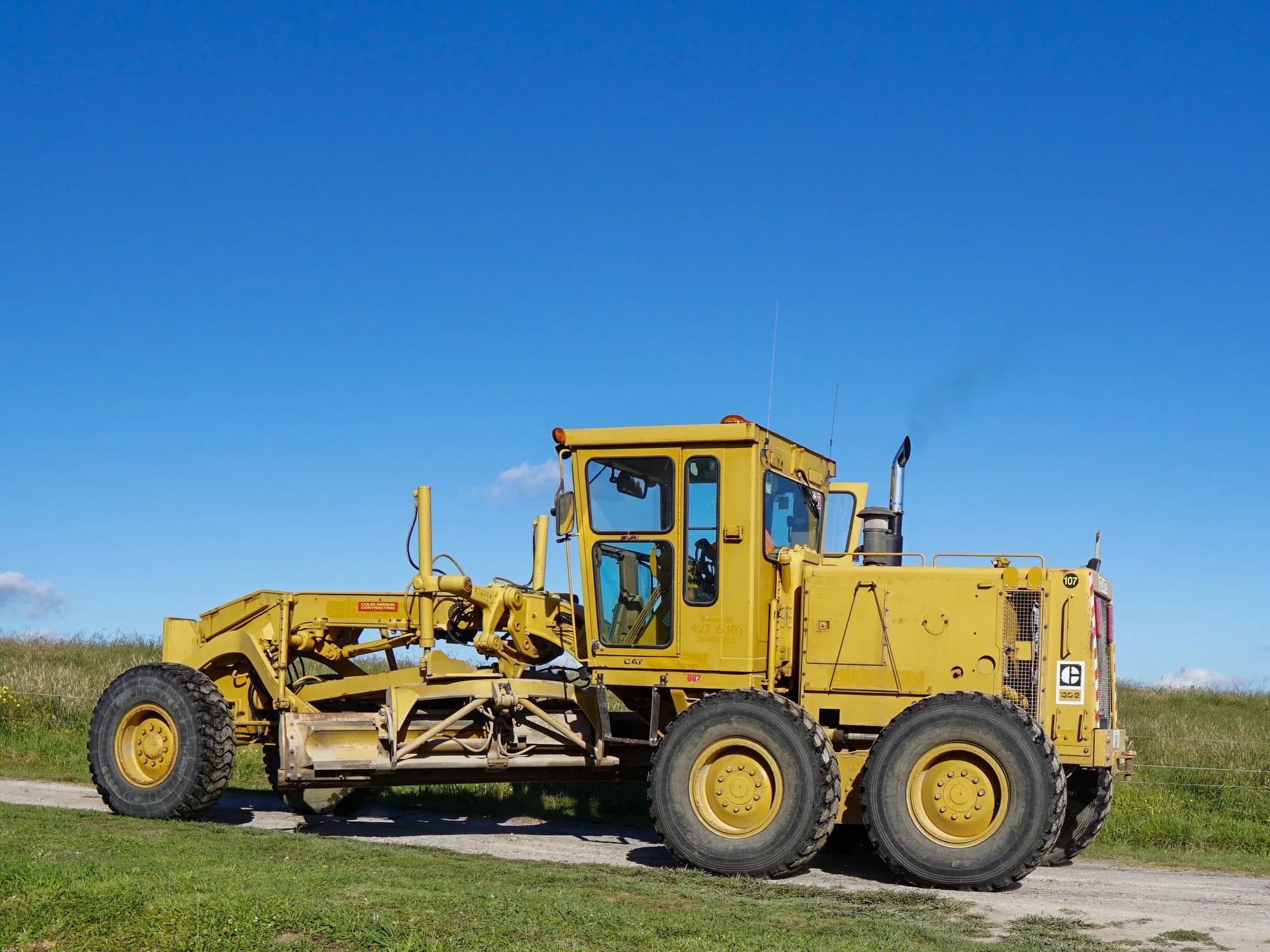 Yellow motor grader working on a rural road beneath a blue sky.